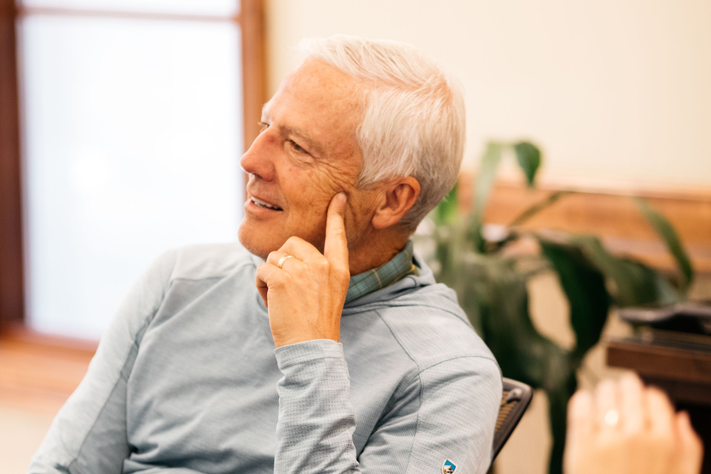 A close-up of a man with short hair, smiling and resting his index finger on his cheek. He is wearing a light blue long-sleeve top with a collared shirt underneath. The background shows a softly focused indoor setting with a large leafy plant and wooden trim along the wall.