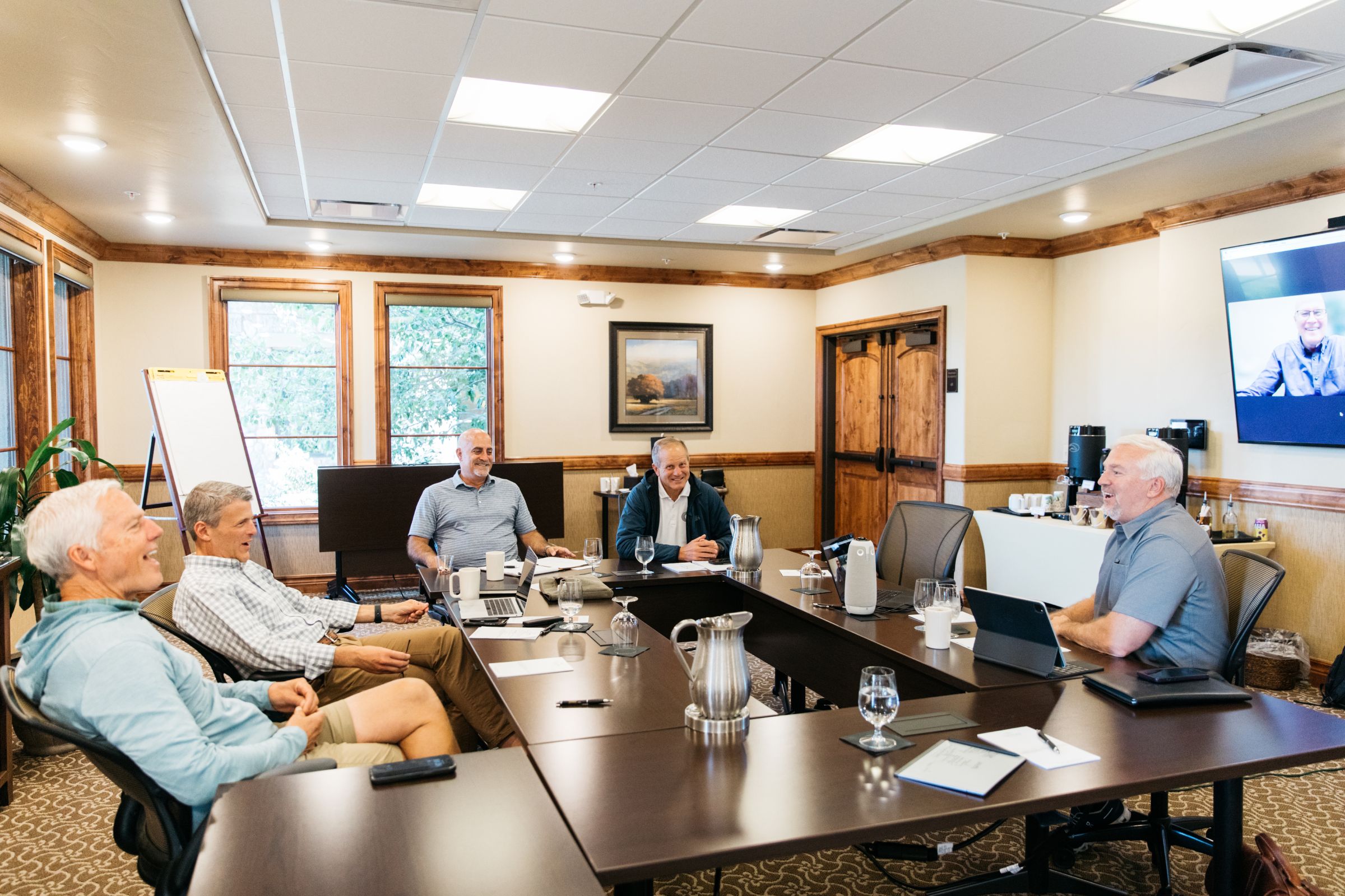 A group of six men in a conference room engaged in a meeting. Five men are seated around a large U-shaped table, while a sixth man participates via video call on a screen mounted to the wall. The room has wooden trim, large windows, a flipchart, and a coffee station in the corner. The atmosphere appears relaxed and collaborative, with laptops, notepads, water pitchers, and mugs on the table.