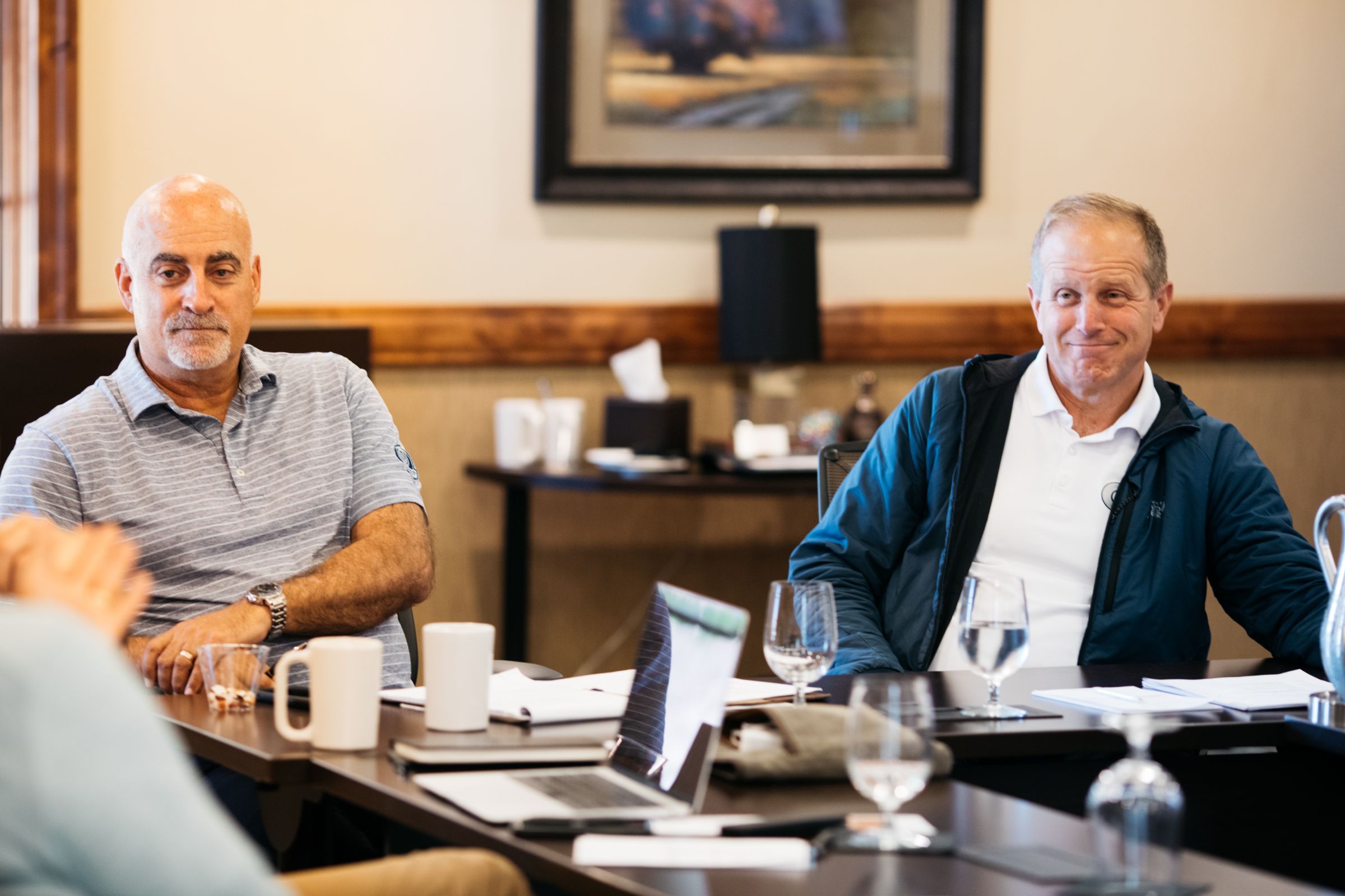 Two men seated at a conference table during a meeting, both appearing engaged and relaxed. The man on the left is wearing a gray polo shirt, while the man on the right is wearing a white polo shirt under a navy blue jacket. Laptops, notebooks, mugs, and glasses of water are on the table in front of them. The background features a wooden panelled wall, a painting, and a small side table with refreshments and supplies.