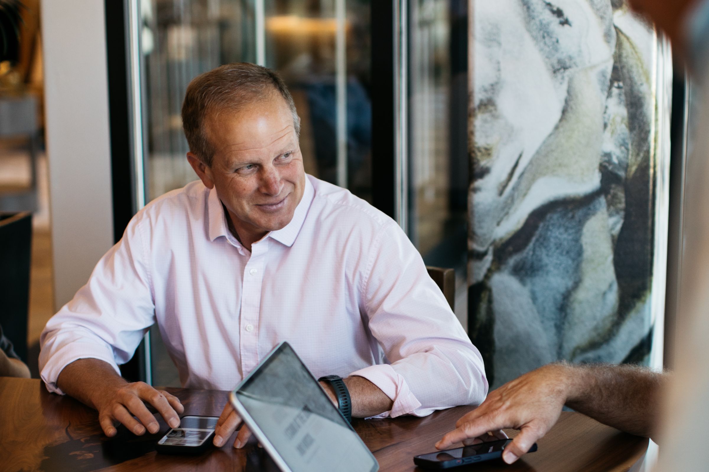 A man is sitting at a table, smiling and engaged in conversation, with his arms resting on the table.