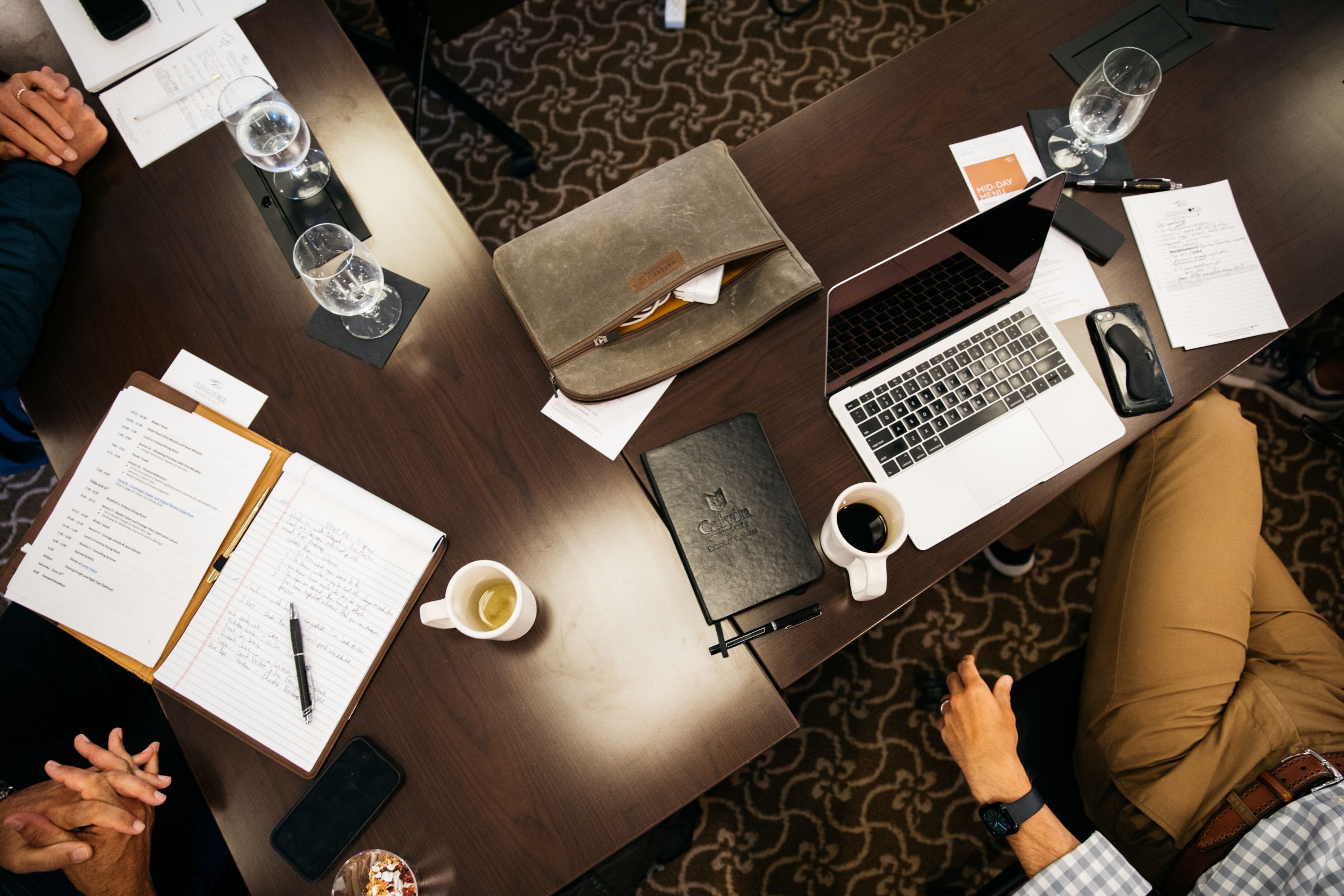 Overhead view of a conference table during a meeting, showing various items including a laptop, notepads with handwritten notes, a leather folder with a printed agenda, a tablet case, a mobile phone, two coffee mugs, several water glasses, and pens.