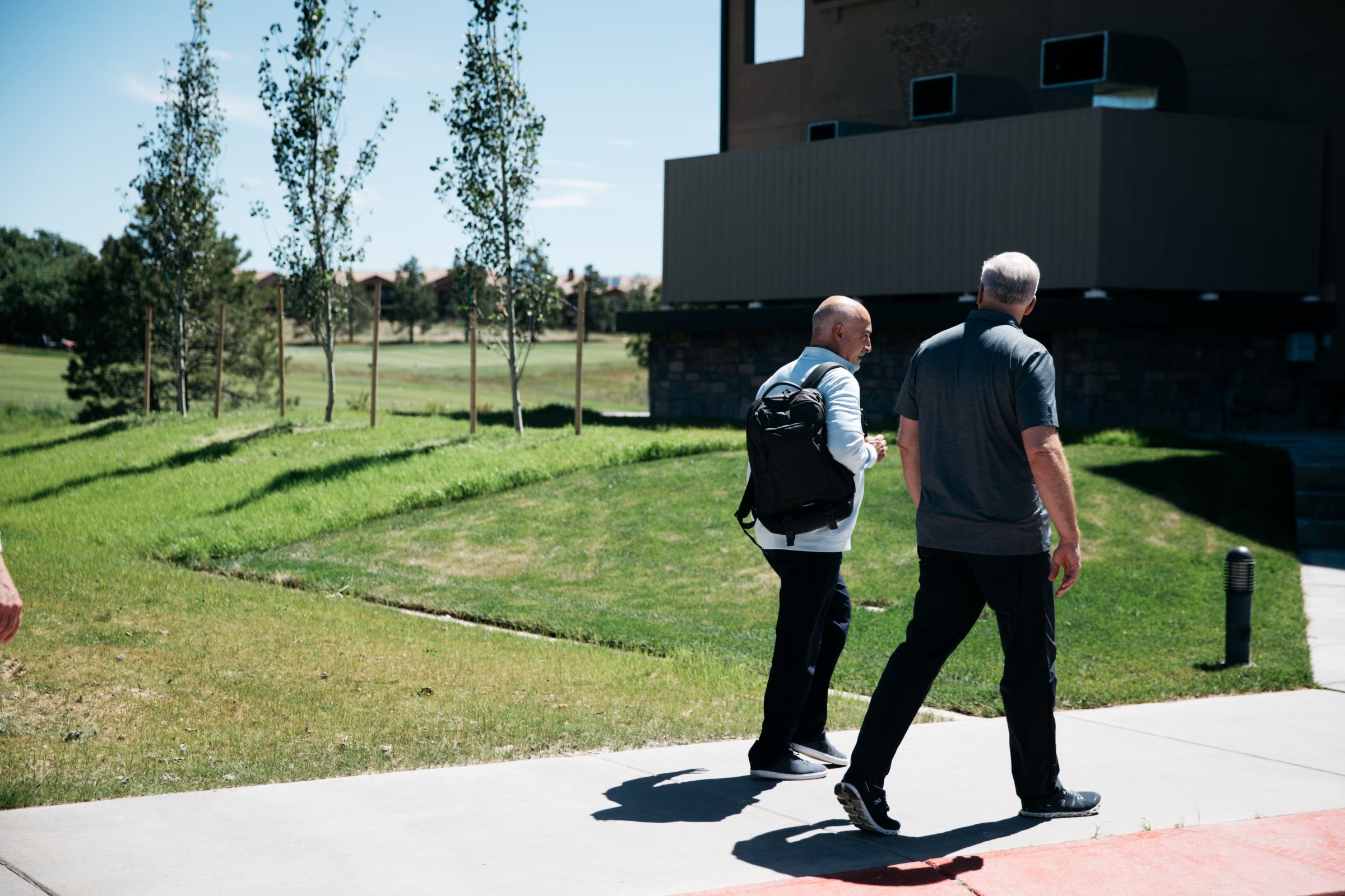 Two men walking on a paved path beside a grassy area, engaged in conversation. One man is wearing a white top and carrying a black backpack, while the other wears a dark grey polo shirt. They are near a modern building, and the background includes young trees, a lawn, and a glimpse of open park land.