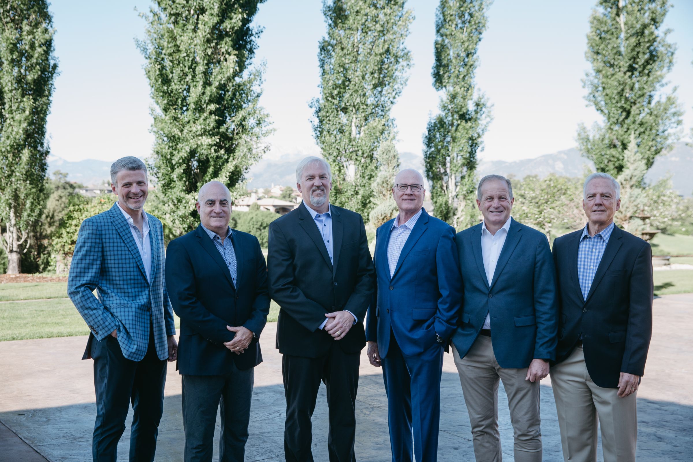 A group of six men standing outdoors in front of tall trees and distant mountains, posing for a photo. They are dressed in business attire, mostly wearing blazers, button-down shirts, and trousers. The setting is bright and clear, suggesting a formal gathering or professional event in a scenic location.